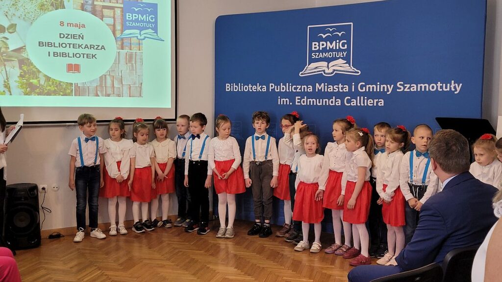 Grupa przedszkolaków ubranych na biało-czerwono stoi przed ścianką z napisem Biblioteka Publiczna Miasta i Gminy Szamotuły.