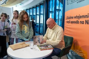 Wnętrze biblioteki. Na pierwszym planie stół z książkami. Z prawej strony siedzi na fotelu aktor Cezary Harasimowicz. Aktor umieszcza autografy na książkach. Obok stołu stoją dzieci.