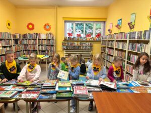 Wnętrze biblioteki. Na pierwszym planie stoją stoły, na których leżą książki. Dalej stoją dzieci i przeglądają książki. Na dalszym palnie na dywanie siedzą dzieci, które oglądają ksiązki
