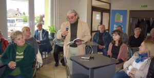 Mężczyzna w jasnej marynarce, jasnych spodniach i ciemnym swetrze, ma na oczach okulary i czyta wiersz poetki. W tle publiczność słucha z uwagą prezentowanych wierszy.