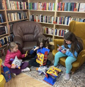 Trzy dziewczynki bawią się w kąciku bibliotecznym z fotelami i dywanem. Dwie trzymają poduszki w kształcie sów. Jedna otwiera pudełko. W tle regały.