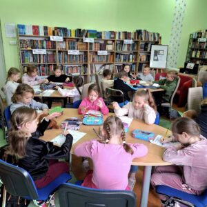 Dzieci klasy drugiej w szkolnej bibliotece. Siedzą przy stolikach po 5-6 osób. Wykonują zadania czytelnicze na kartach pracy.