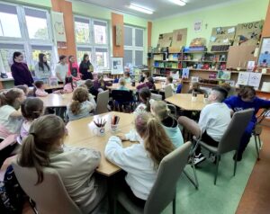 Cztery stoliki w szkolnej bibliotece, przy których siedzą dzieci klas 1-3 (około 23 osób), dzieci rozmawiają ze starszymi koleżankami (5 dziewcząt), które stoją przed nimi przy oknach i prezentują swoje książki z dzieciństwa.