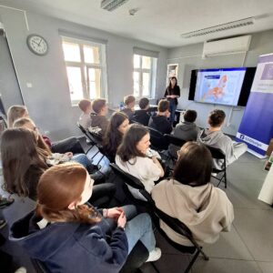 Uczniowie klasy 7 siedzący w rzędach w bibliotece, przed nimi osoba prowadząca spotkanie, w tle prezentacja multimedialna z mapą Europy