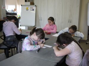 Grupa dzieci rysuje przy stole w sali bibliotecznej, skupione na twórczym zadaniu.
