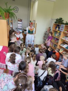 Grupa dzieci w bibliotece szkolnej patrzy na kobietę w tradycyjnym stroju śląskim.