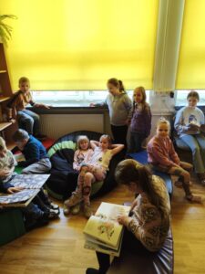 Dzieci siedzą na pufach, czytają książki. Pomieszczenie biblioteki ma przesłoniete żółtymi roletami okna.