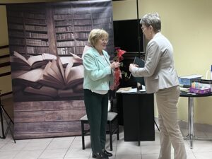Dwie kobiety ubrane odświętnie. jedna wręcza drugiej różę i dyplom.