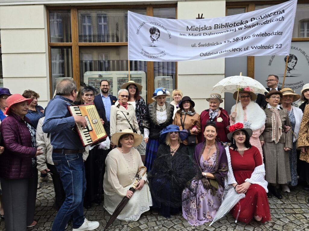 Grupa osób ubranych w eleganckie stroje stylizowane na początek 20. wieku pozuje do zdjęcia. Trzymają transparent z nazwą biblioteki.