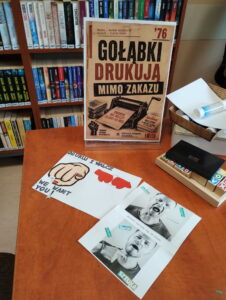 Kartki z wydrukowanymi zdjęciami i grafiką, leżące na stole. Obok stoi plakat w stojaku z pleksiglasu.