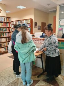 Trzy kobiety stoją przy stoliku. Pochylają się nad czymś. W tle regały biblioteczne.