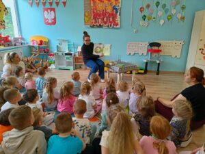 Sala przedszkolna. Nauczycielka siedzi na krześle i czyta dzieciom książkę, pokazując ilustracje. Grupa kilkunastu dzieci siedzi na dywanie i uważnie słucha.