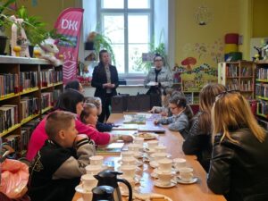 Przy długim stole siedzą dzieci i dorośli, na stole stoją filiżanki, talerzyki z ciasteczkami oraz leżą książki. W tle dwie kobiety stoją przed grupą i prowadzą spotkanie. 