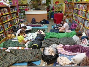 Między regałami pełnymi książek na podłodze rozłożone są śpiwory, karimaty, poduszki i plecaki. Kilkoro dzieci siedzi lub leży, czytając książki i czasopisma. 