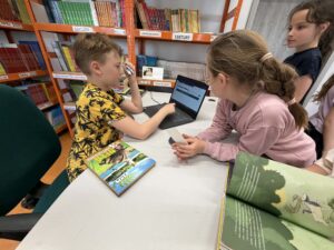 Chłopiec wypożycza książkę dziewczynce przy stanowisku bibliotekarza. Ma przed sobą komputer.