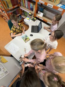 Przy stanowisku bibliotekarza siedzi chłopiec. Grupa uczniów wybiera położone na biurku zakładki do książek.