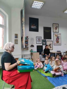 Prowadząca trzyma na kolanach walizkę z rekwizytami teatralnymi. 