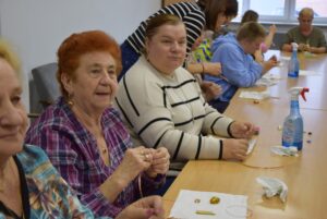 Kobiety w różnym wieku siedzą przy stole, przed sobą mają elementy do tworzenia biżuterii.